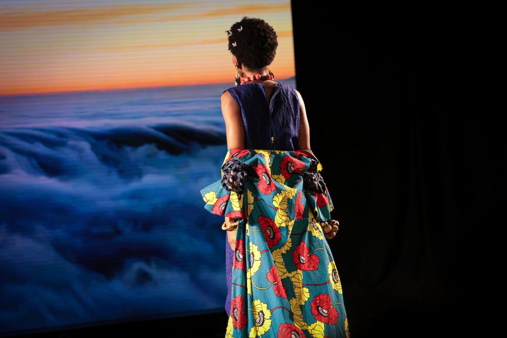 Fashion model wearing colorful floral dress against sunset backdrop on runway.