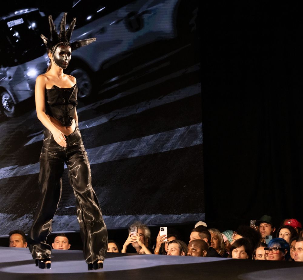 A model wearing an avant-garde black outfit struts down a dramatically lit catwalk.