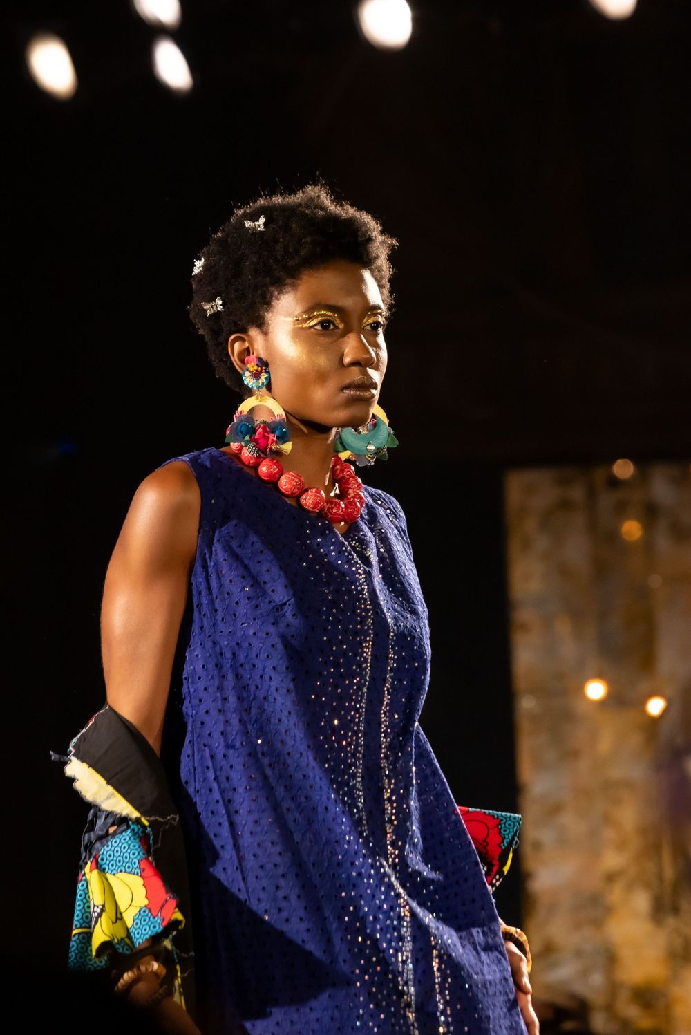 Model showcasing navy dress with red beaded necklace and colorful accessories on catwalk.