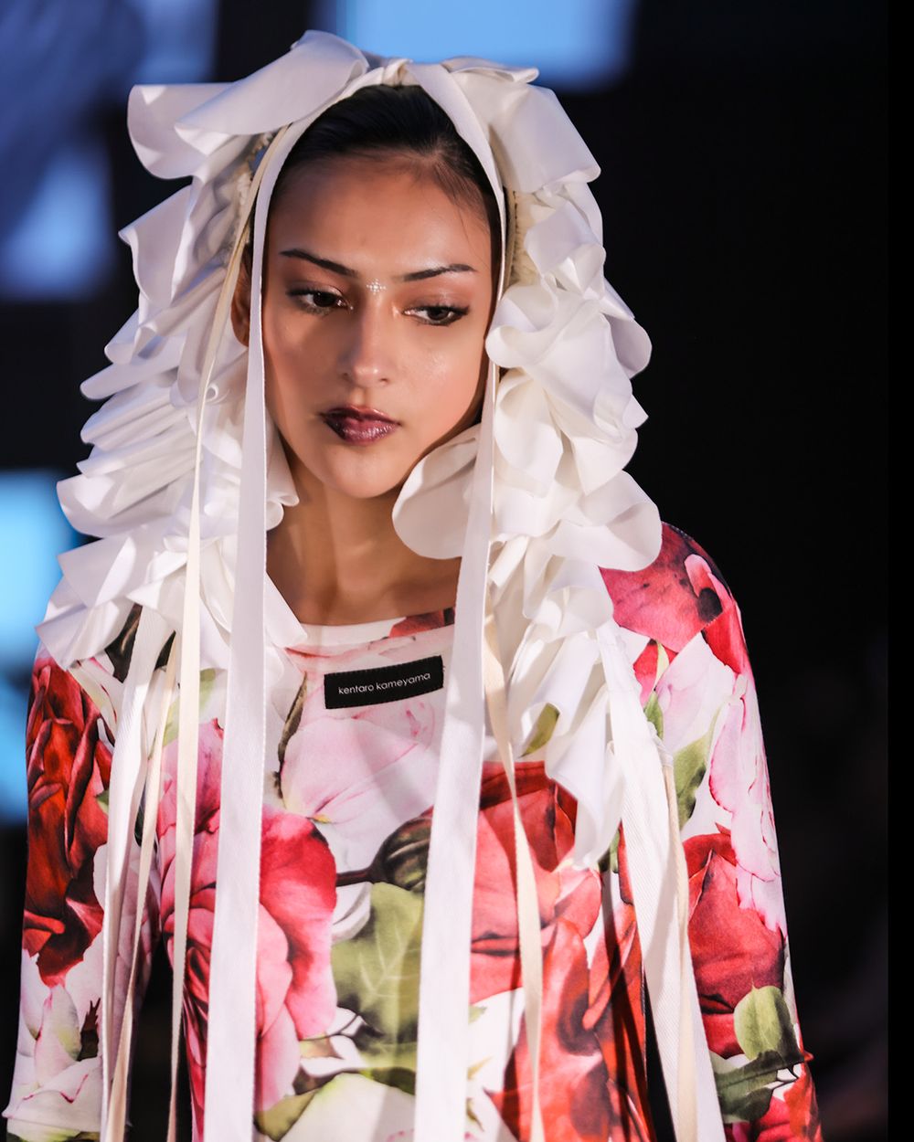 Fashion model wearing a floral print dress with white ruffled headpiece walks runway during catwalk show.