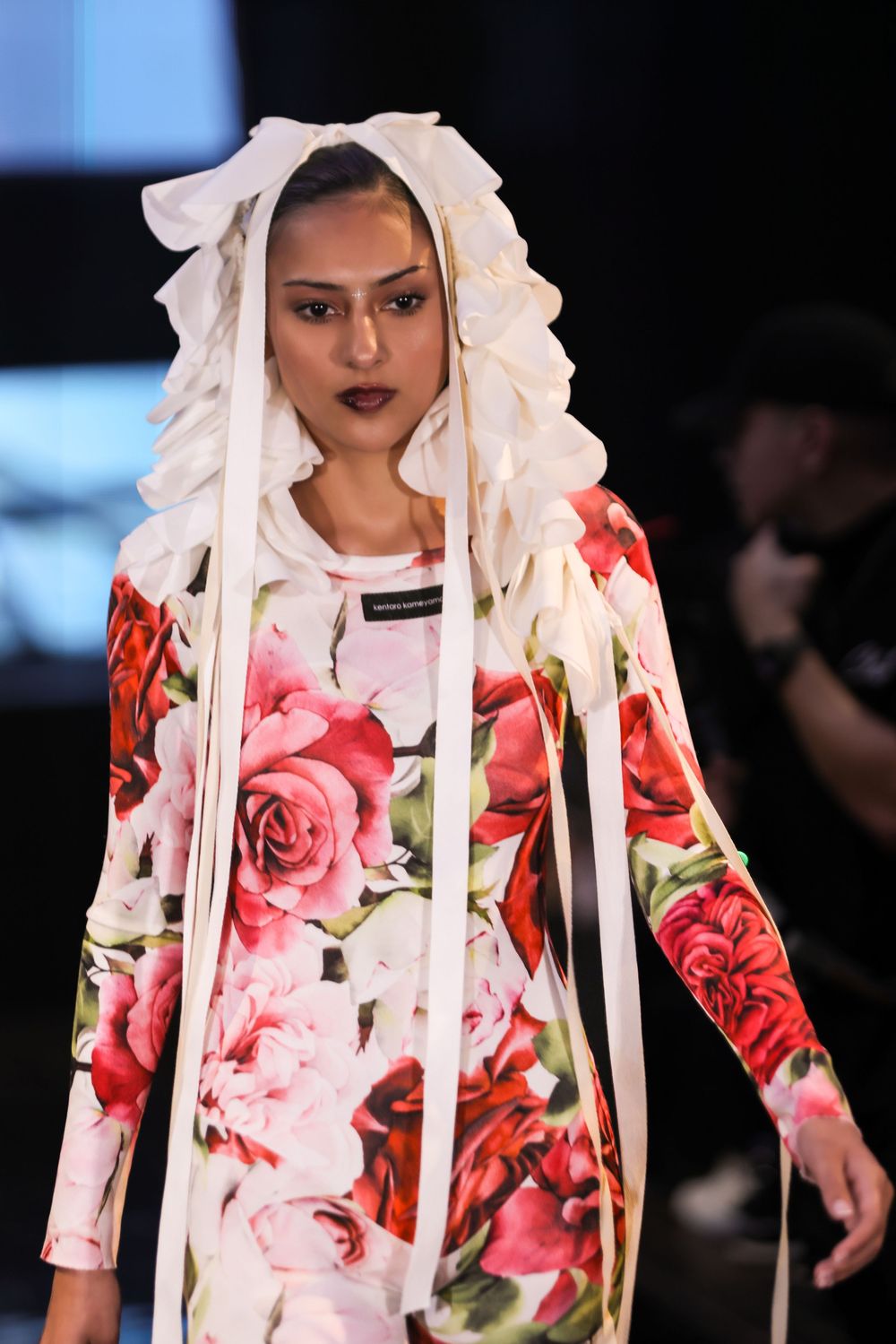 Model walks runway in floral print dress with white head covering during fashion show.
