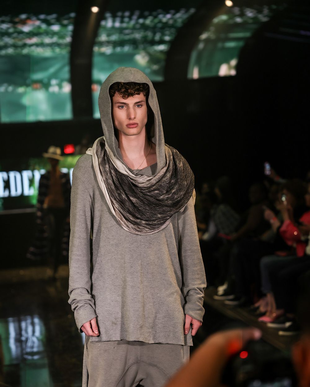 Fashion model on catwalk wearing loose grey top with draped cowl neckline during runway show presentation.