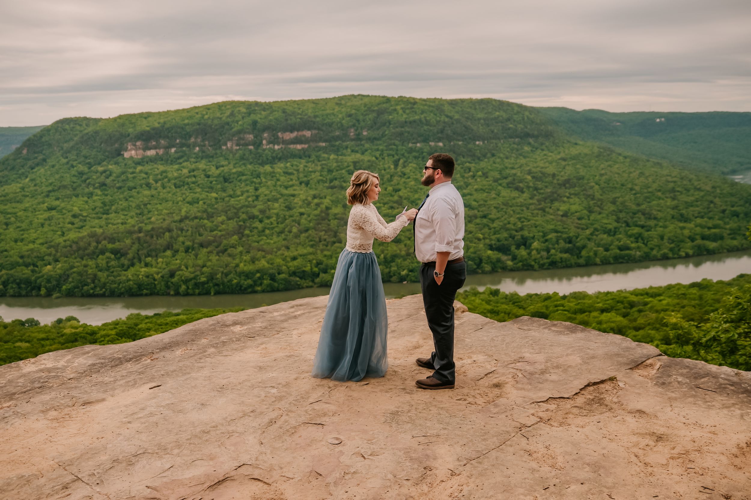 A scenic mountain overlook with green forested hills stretching into the distance under cloudy skies.