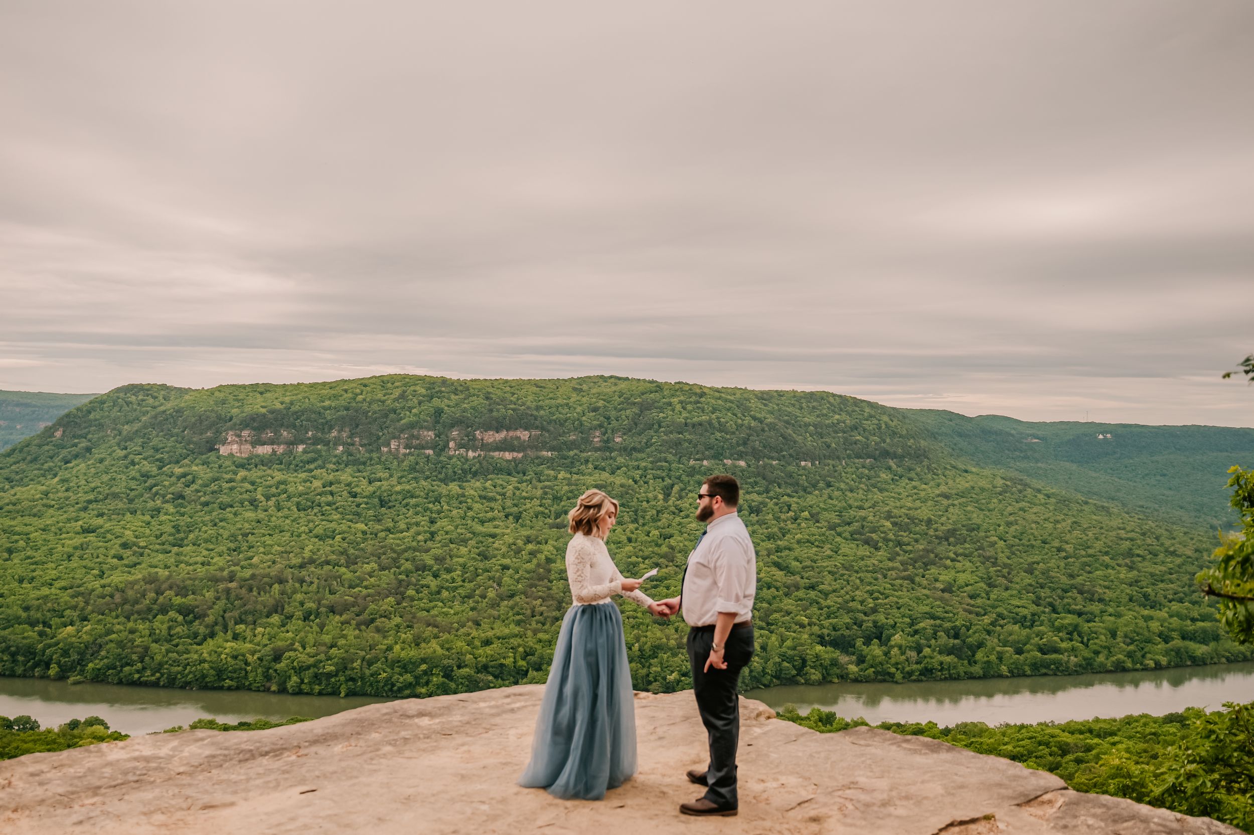 Two people stand on a rocky mountain overlook with sweeping valley views below.
