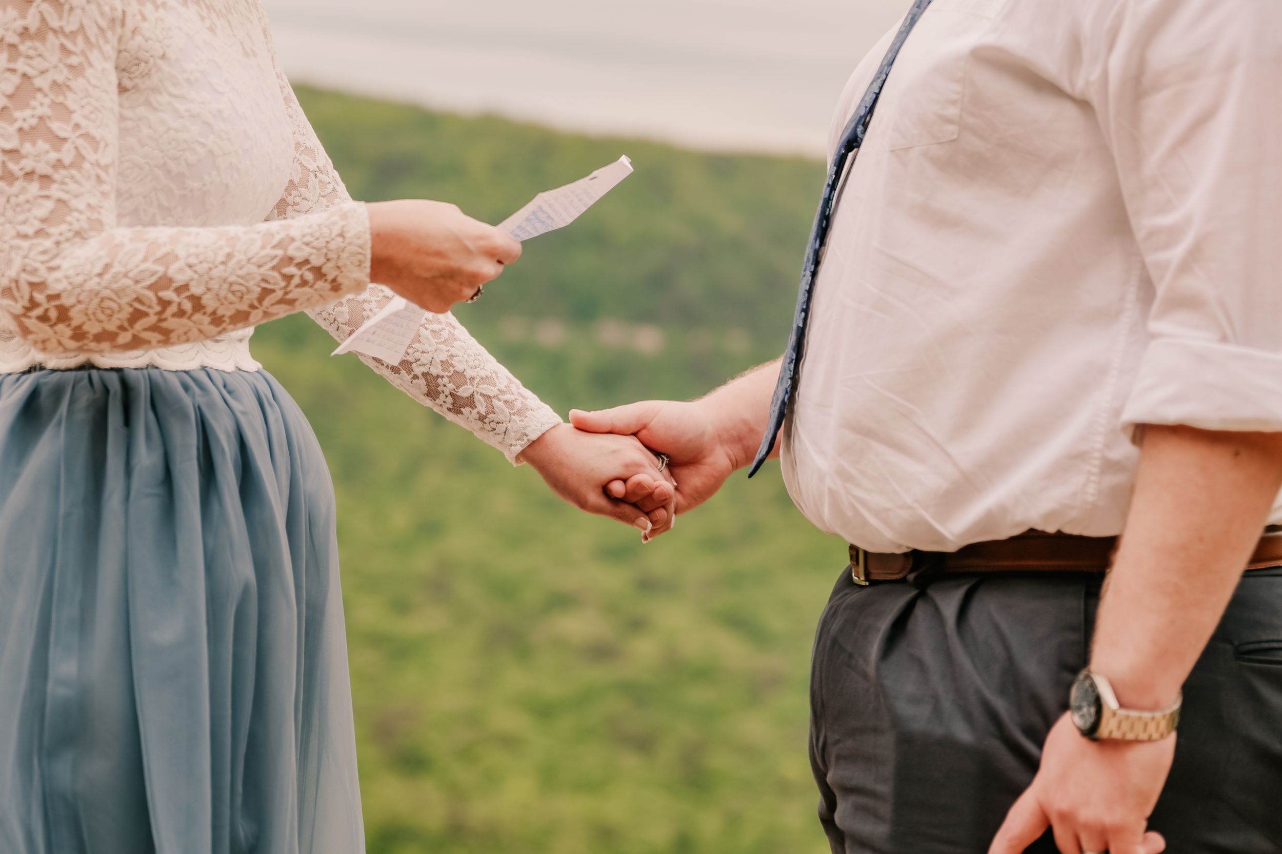 A couple holds hands against a backdrop of rolling green mountains and cloudy skies.