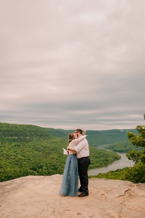 A couple embraces on a scenic overlook with rolling green mountains and a winding river in the background.