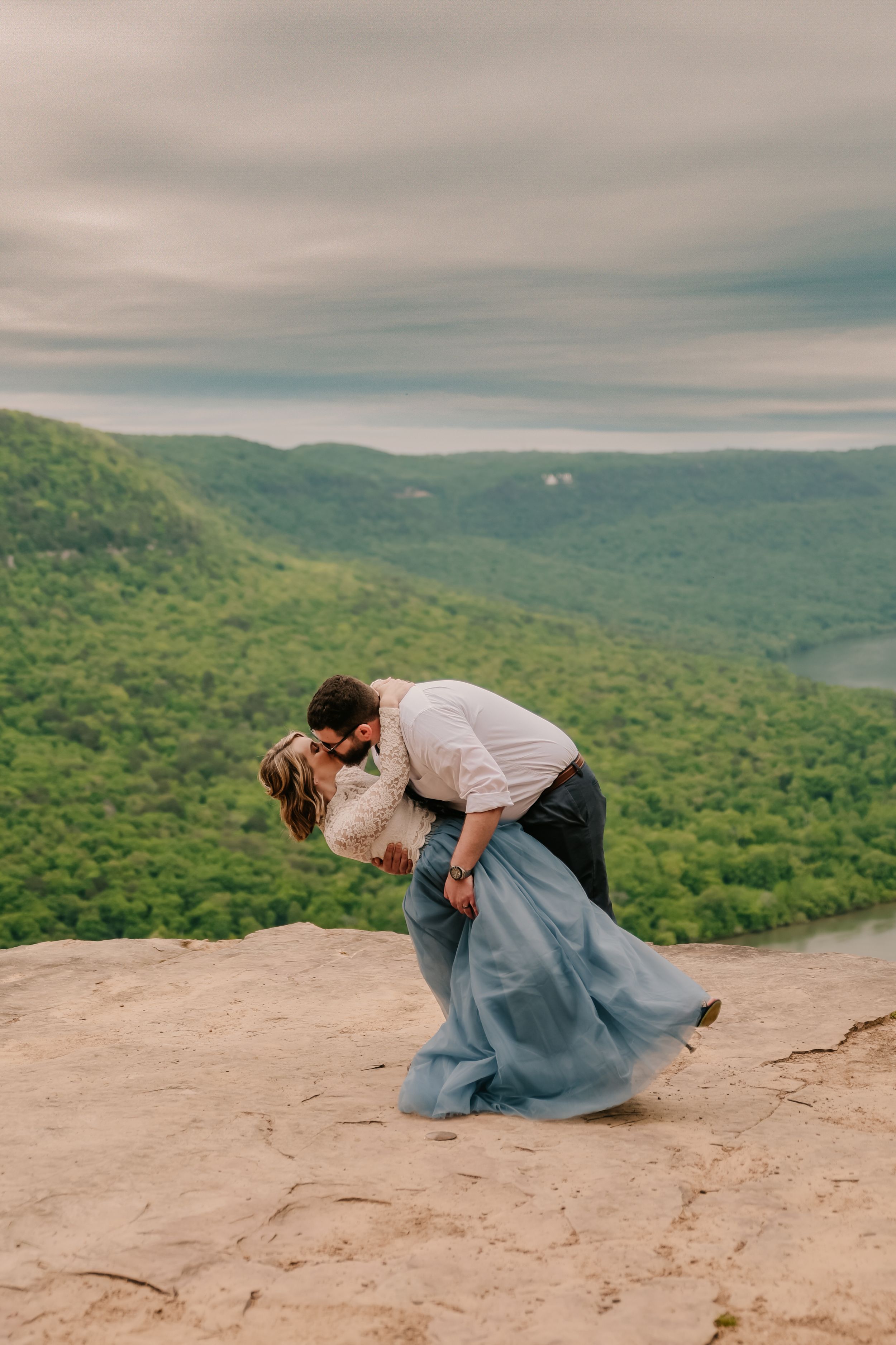 A romantic dip pose photographed on a rocky mountain ledge overlooking a green valley.