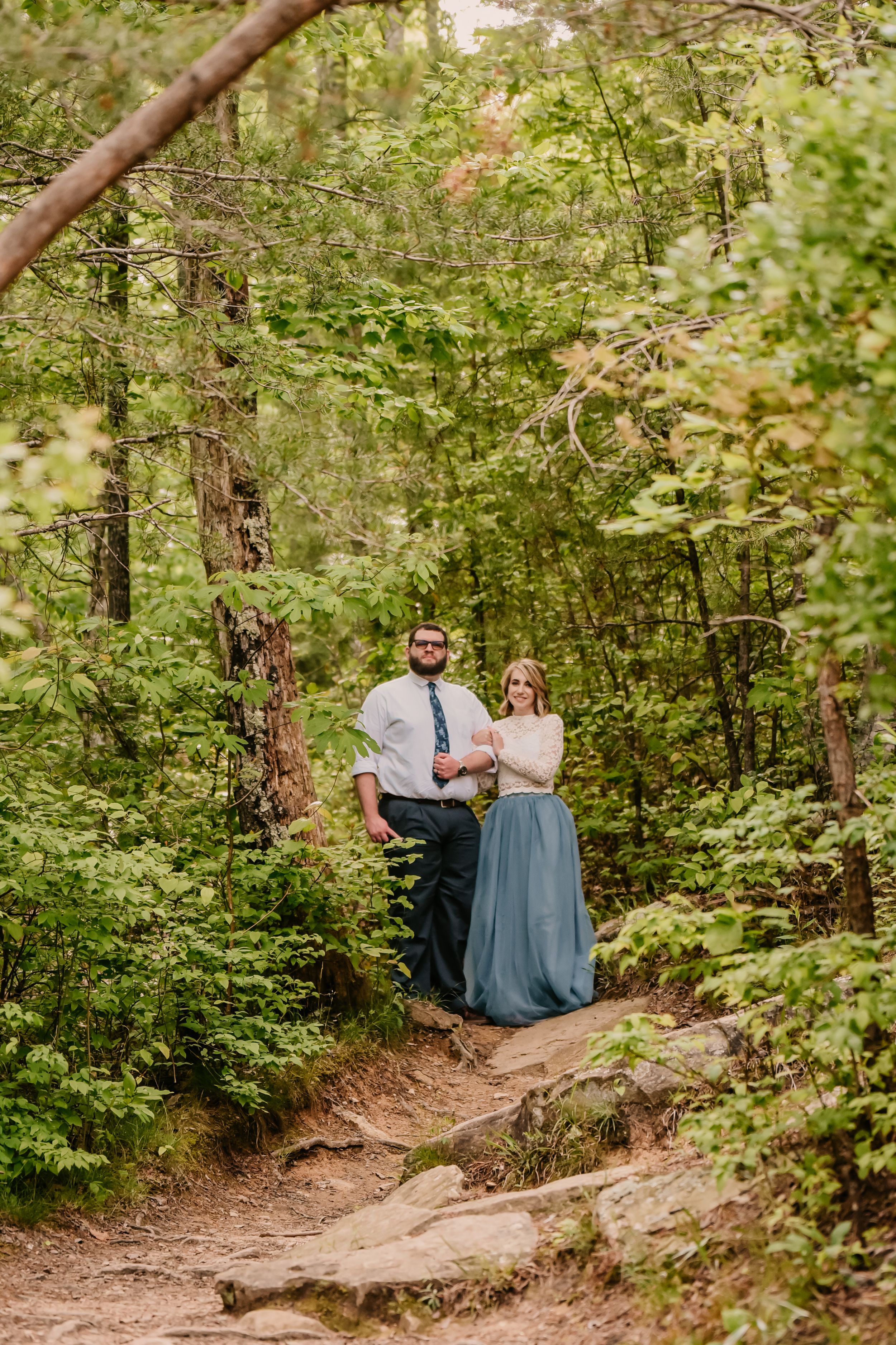 A couple walks hand in hand along a wooded forest trail surrounded by green foliage.