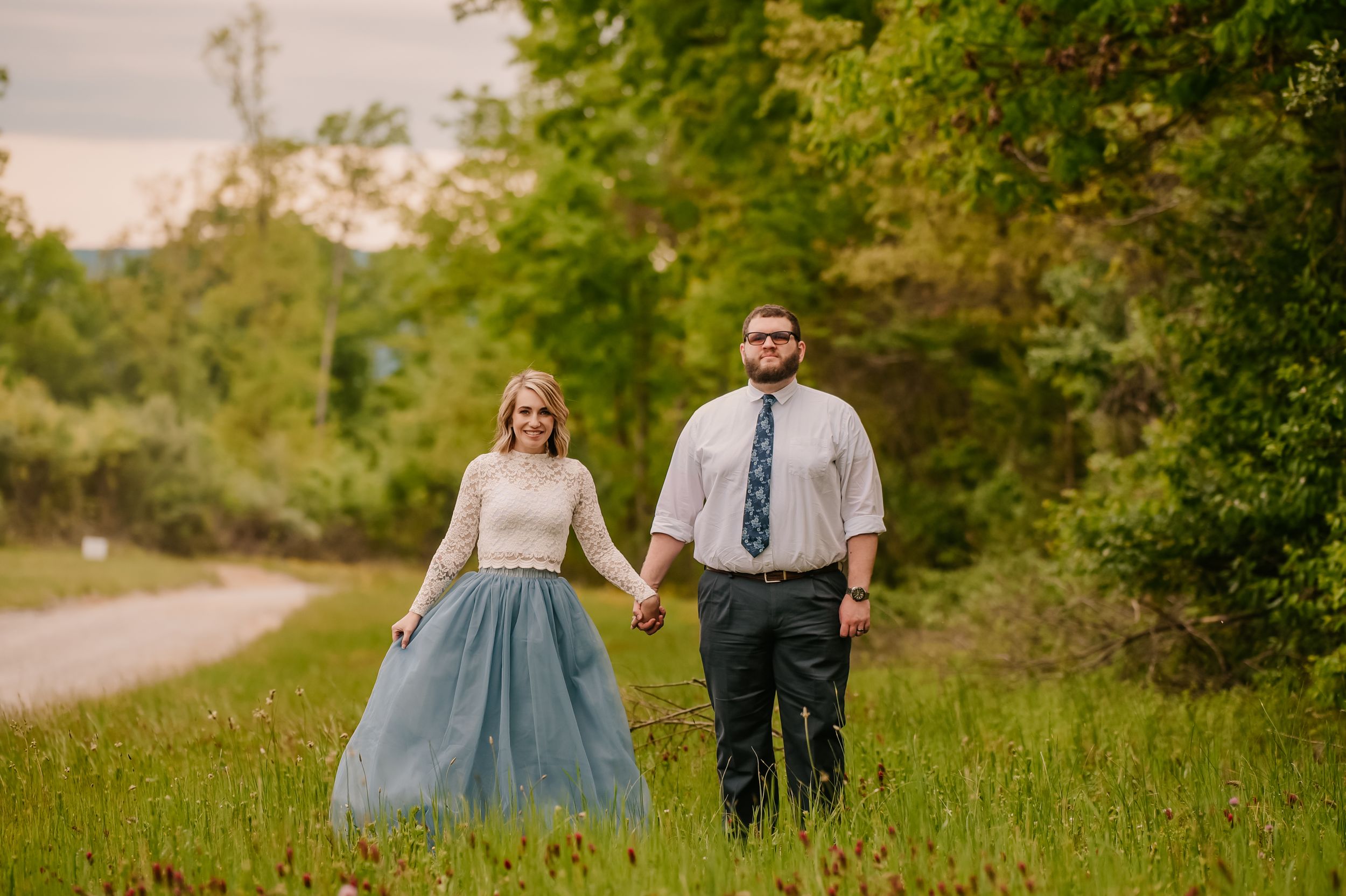 A couple strolls through a sunny meadow surrounded by trees and natural greenery.