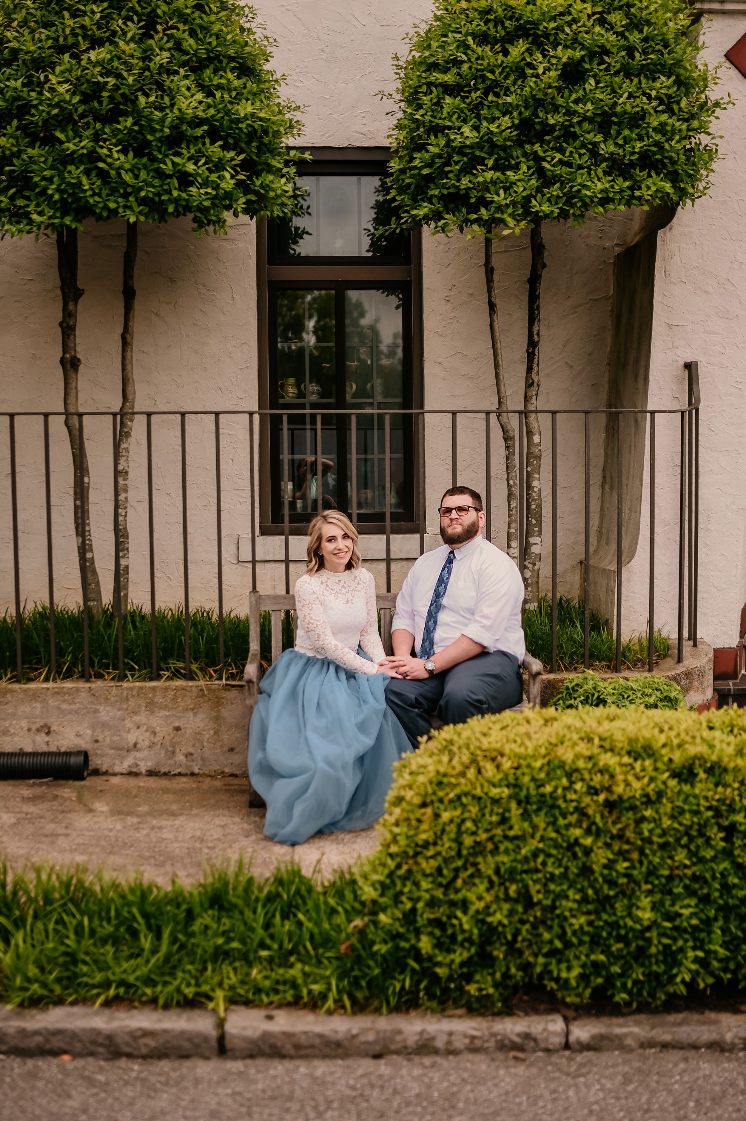 A couple sits on steps of an elegant building with manicured hedges and symmetrical trees.