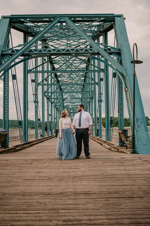 A couple stands on a turquoise steel bridge with intricate architectural details stretching into the distance.