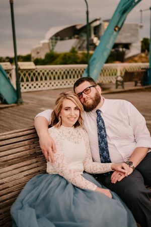 A couple sits together on a wooden bench near a turquoise bridge structure at sunset.