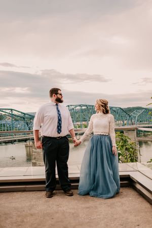A couple holds hands while standing on a platform with a turquoise bridge and river in the background.