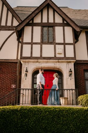 A couple stands on the balcony of a Tudor-style house with a distinctive red door and brick facade.