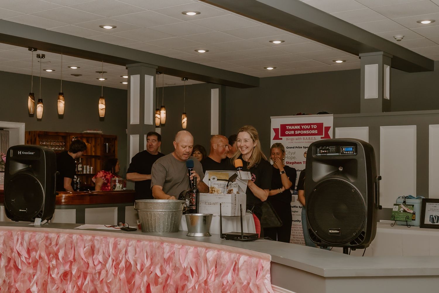 Staff members stand behind a bar counter with audio equipment and pendant lighting in a restaurant setting.
