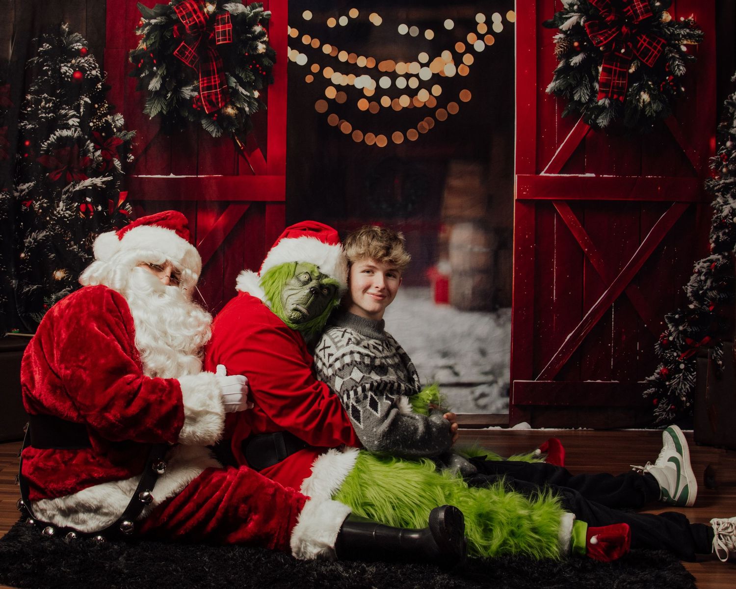 Festive holiday scene with Santa and the Grinch sitting by decorated red doors with string lights.
