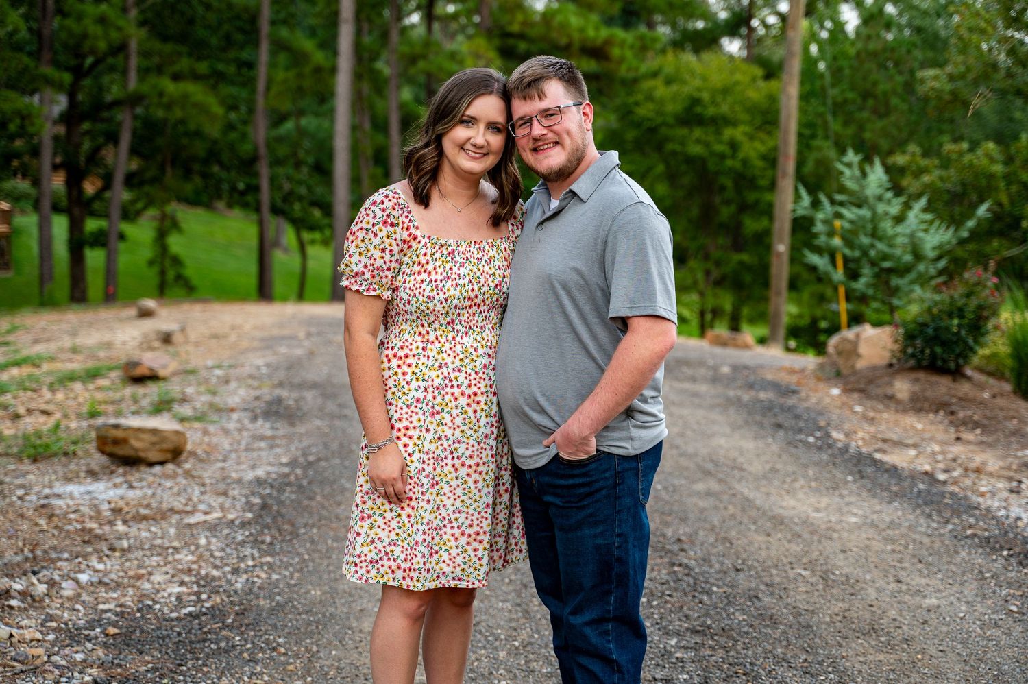 Smiling couple posing together on a gravel path surrounded by lush green trees.