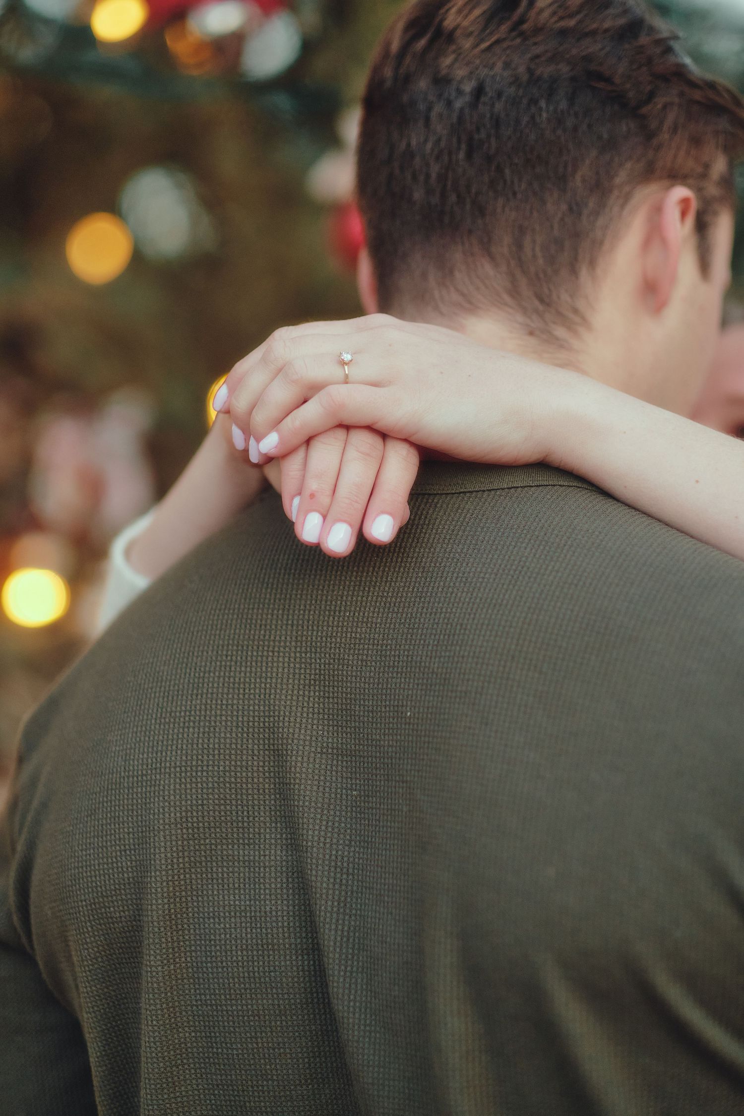 A woman rests her hands on a man's shoulder, engagement ring visible, with warm bokeh lights in the background.