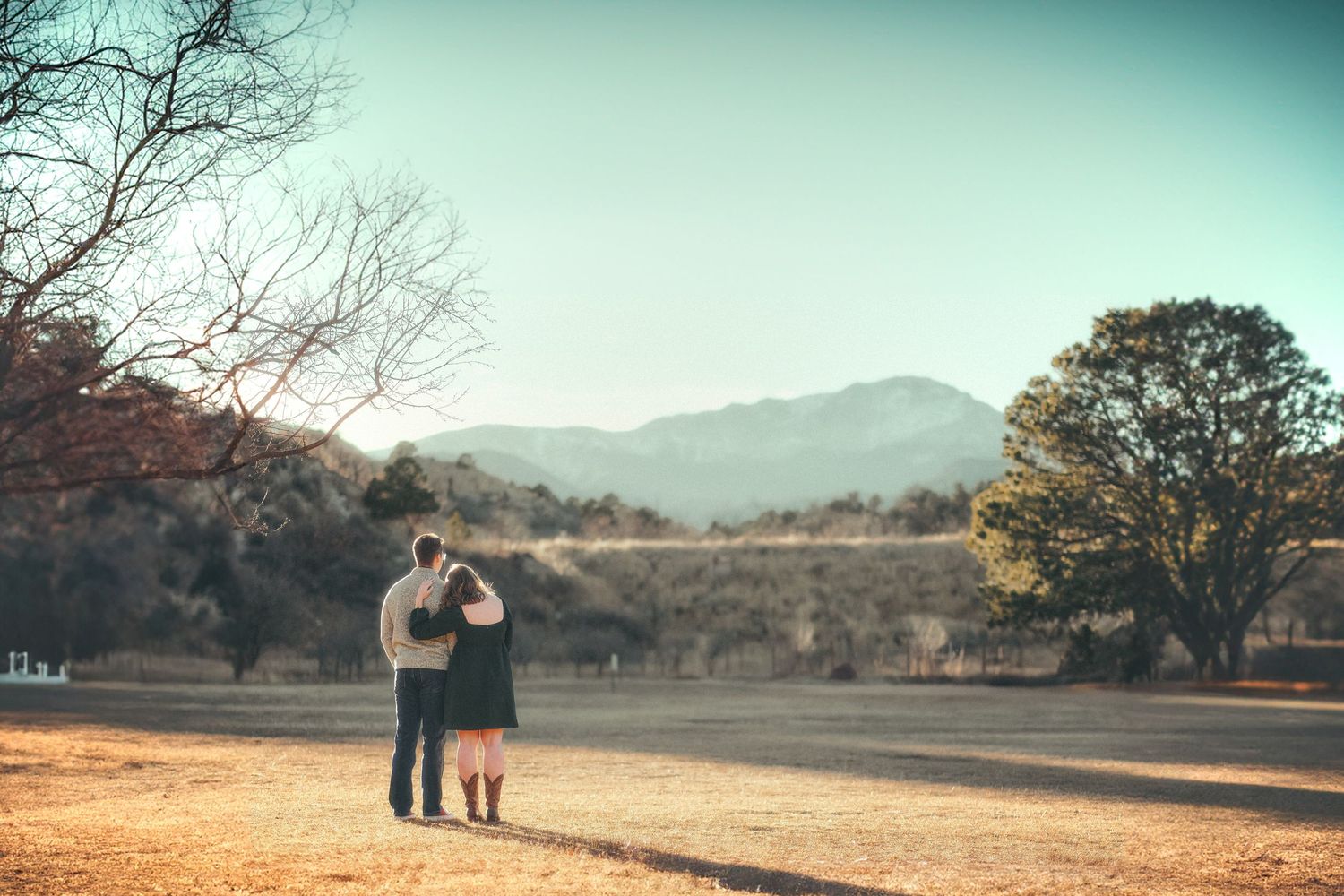 A couple stands in an open field, arms around each other, gazing at distant mountains under a clear sky.
