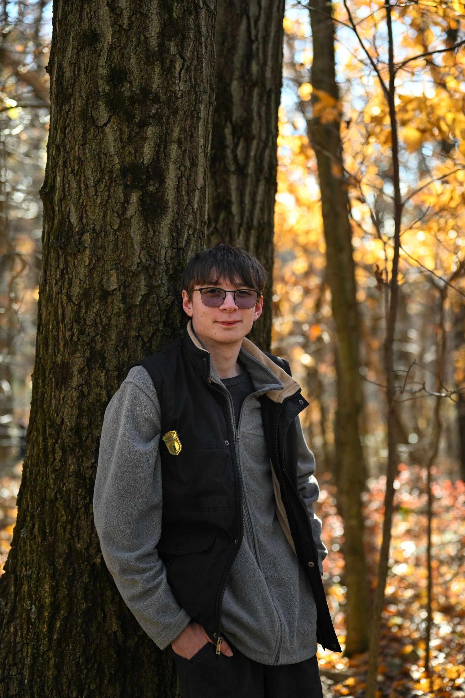 A photograph of a young man smiling and facing the camera leaning up against a tree