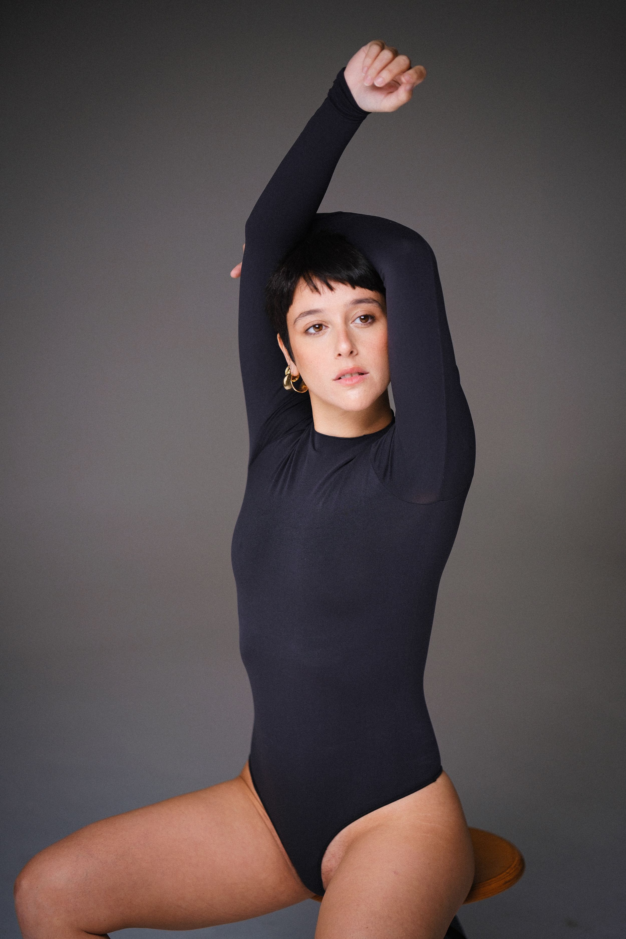 Figure in black long sleeve bodysuit posing on wooden stool against grey backdrop.