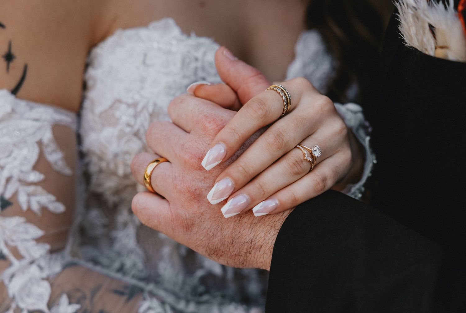 Close-up of newlywed couple's hands showing wedding rings against white lace dress and dark suit.