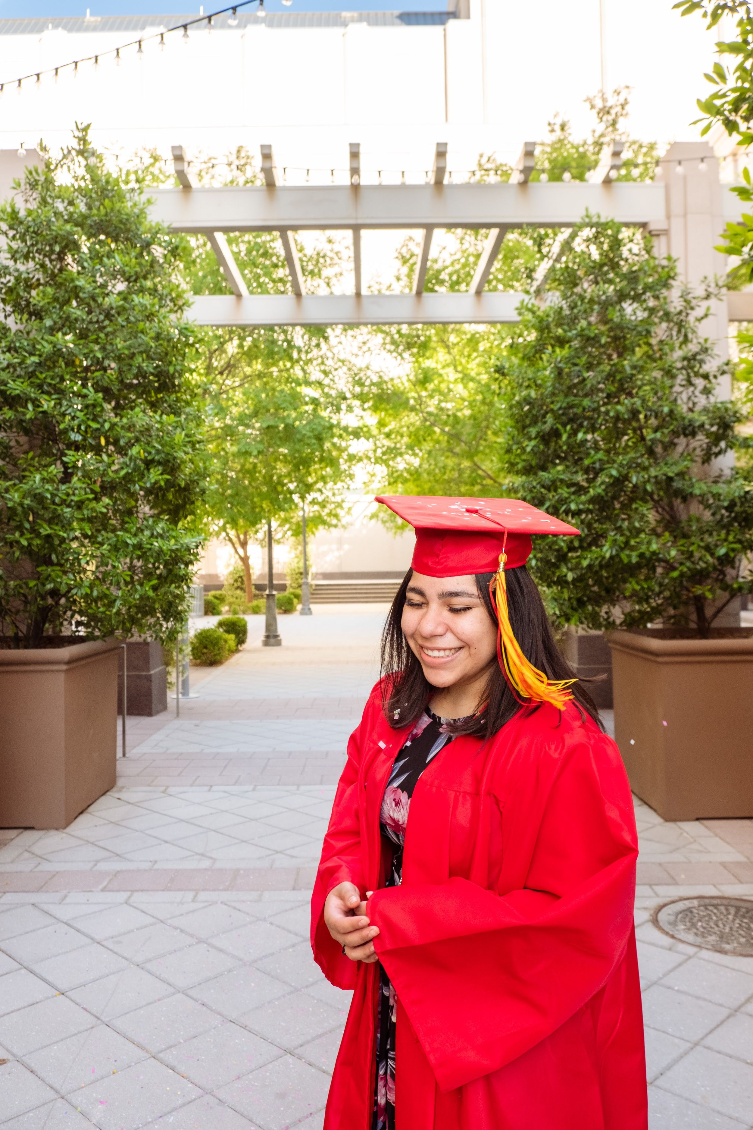 A graduate in red cap and gown smiling in a courtyard with plants.