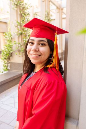 A graduate in bright red academic regalia poses against a light colored wall.