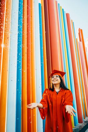 A graduate in red regalia poses joyfully against colorful vertical tubes outdoors.