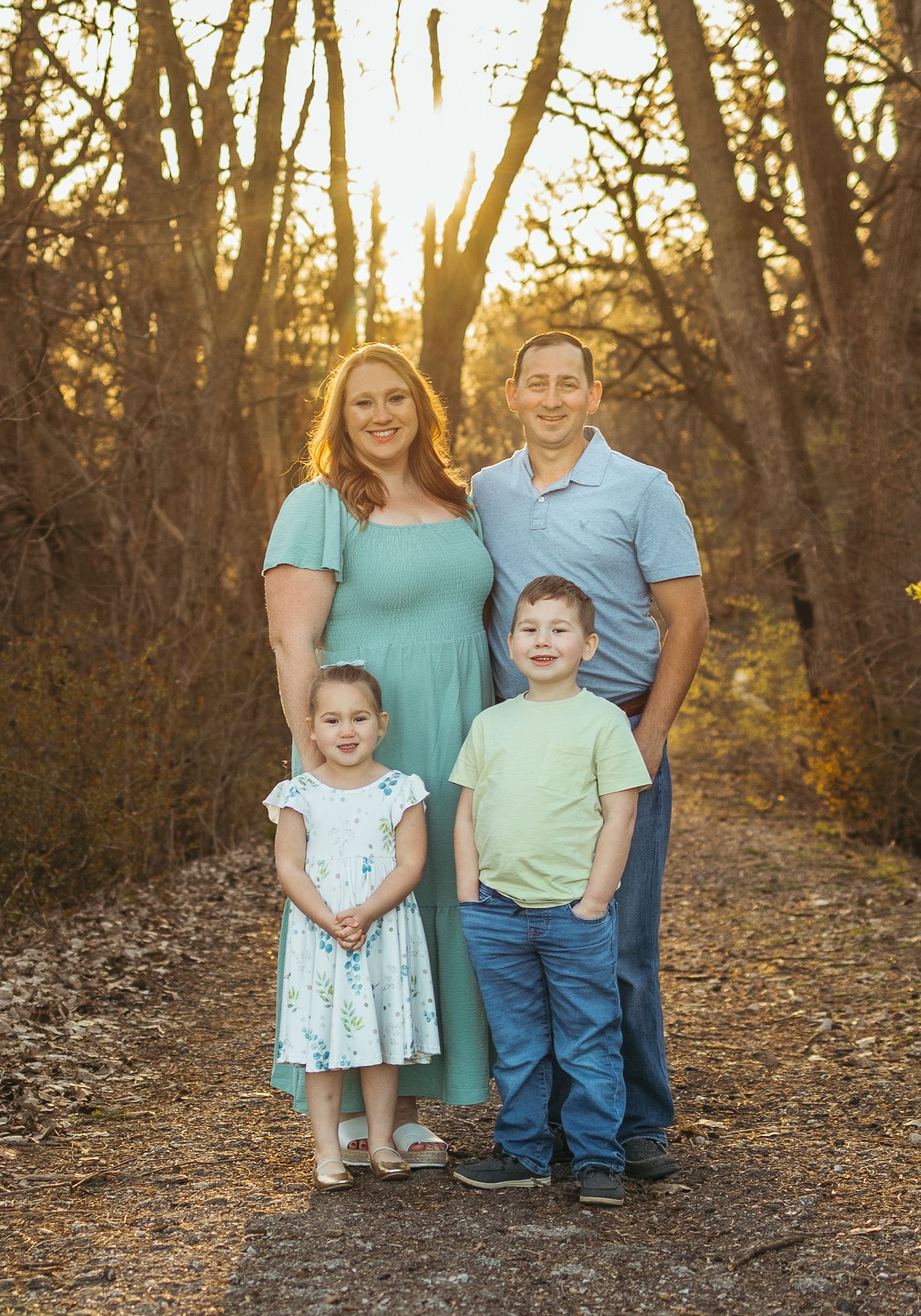 A family poses together on a forest path at sunset with golden light filtering through the trees behind them.
