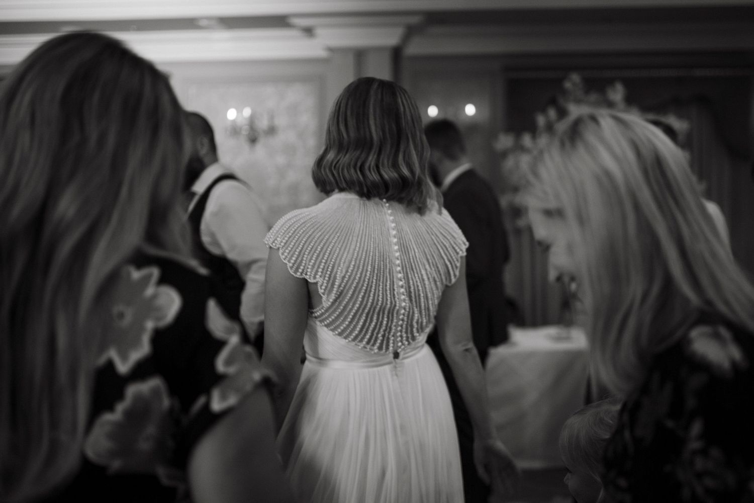 A black and white wedding photo showing a dress with delicate beaded details on the back during a reception celebration.