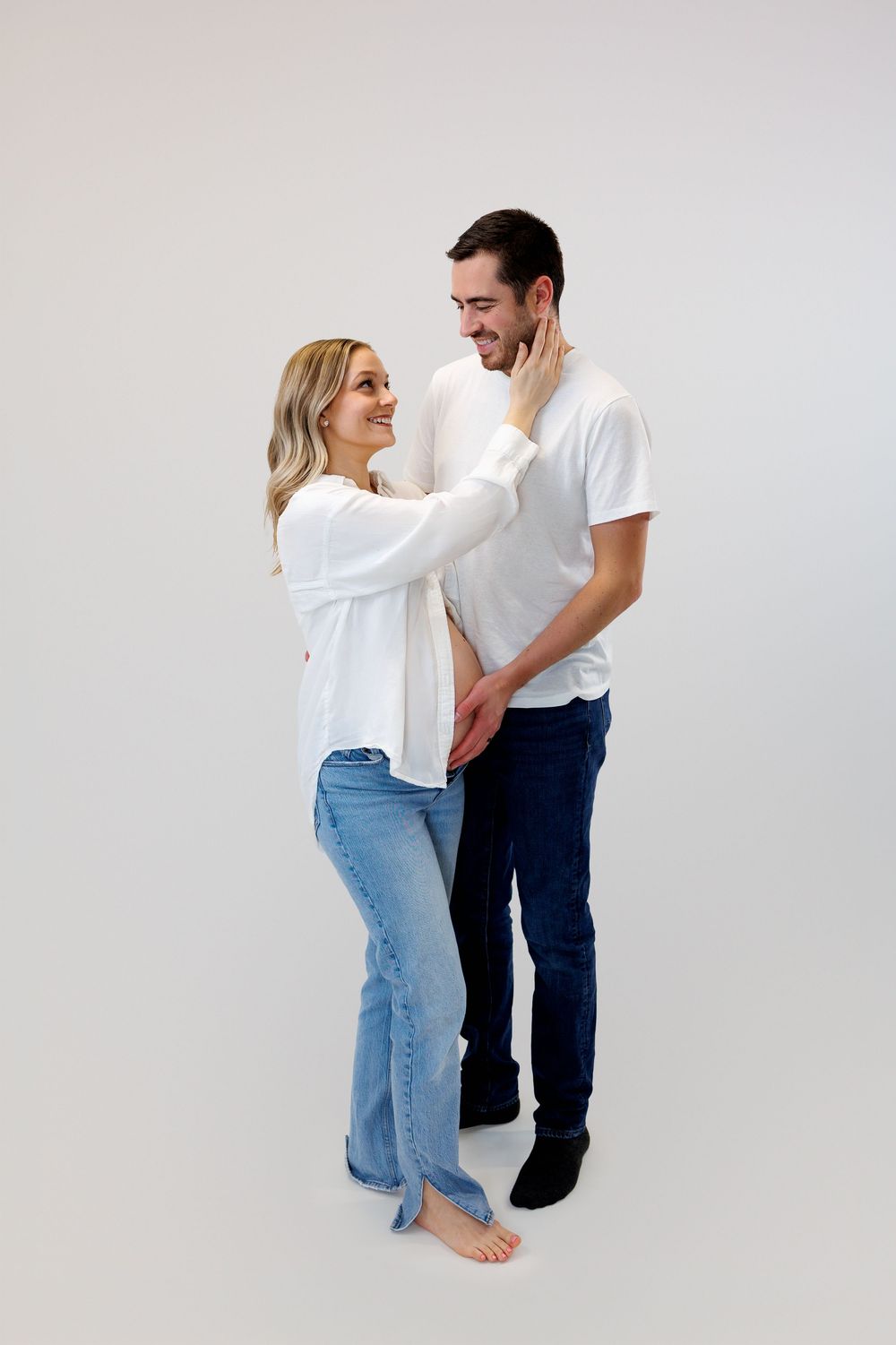 A couple in casual white tops and jeans embraces and dances together against a light gray studio background.