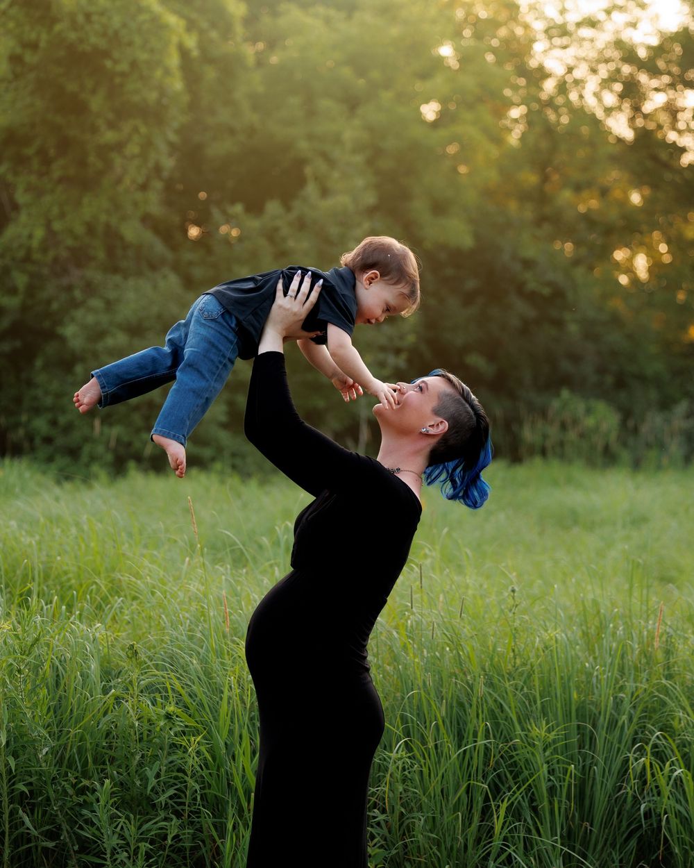 Pregnant woman in black dress lifting toddler overhead in sunny green field, sharing a joyful moment outdoors.