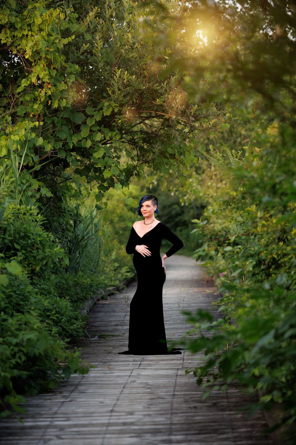 Pregnant woman in elegant black velvet gown standing on wooden path surrounded by lush green trees in golden sunlight.
