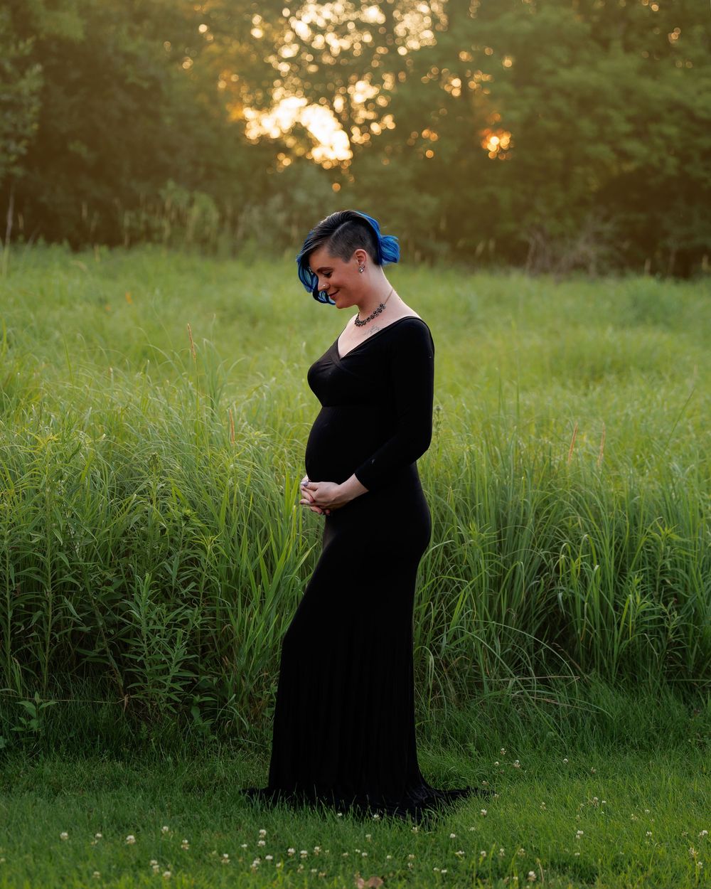 Pregnant woman in black gown standing in green field at golden hour, cradling baby bump with blue hair accessories.