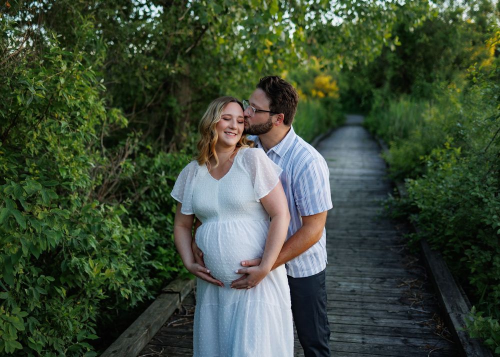 Pregnant couple embracing on a wooden boardwalk path surrounded by lush green trees during golden hour.