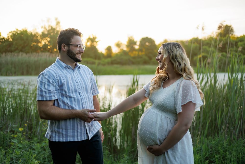 Pregnant couple holding hands outdoors near a lake at golden hour, woman in white dress cradling baby bump.