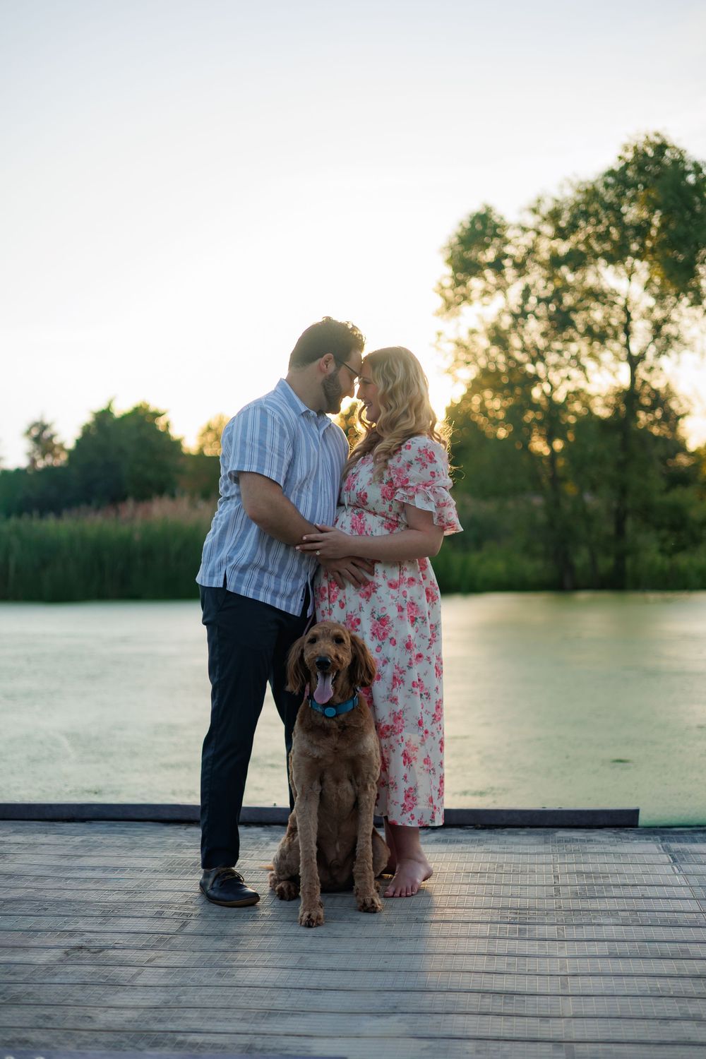 Couple sharing a romantic moment on a dock by a lake at sunset, with their golden retriever dog sitting beside them.