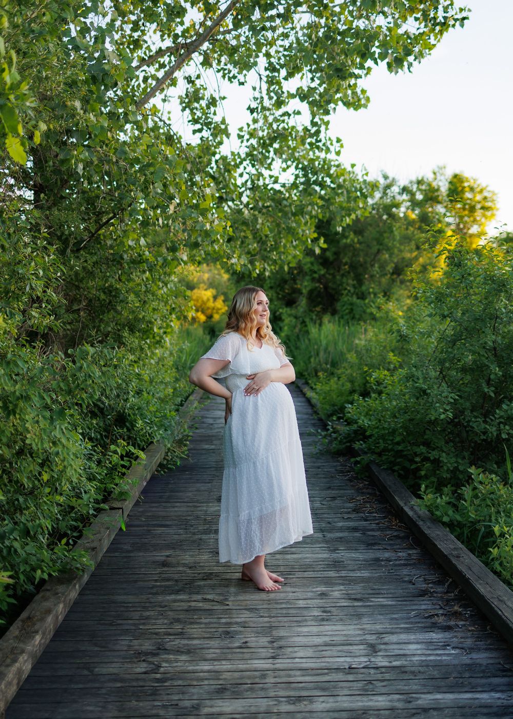 Pregnant woman in white dress standing on wooden boardwalk surrounded by lush green trees during golden hour.