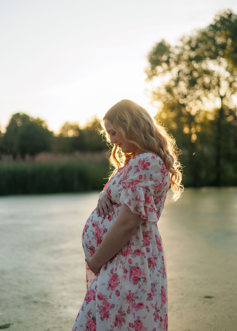 Pregnant woman in floral dress holding baby bump by a sunlit lake at golden hour.
