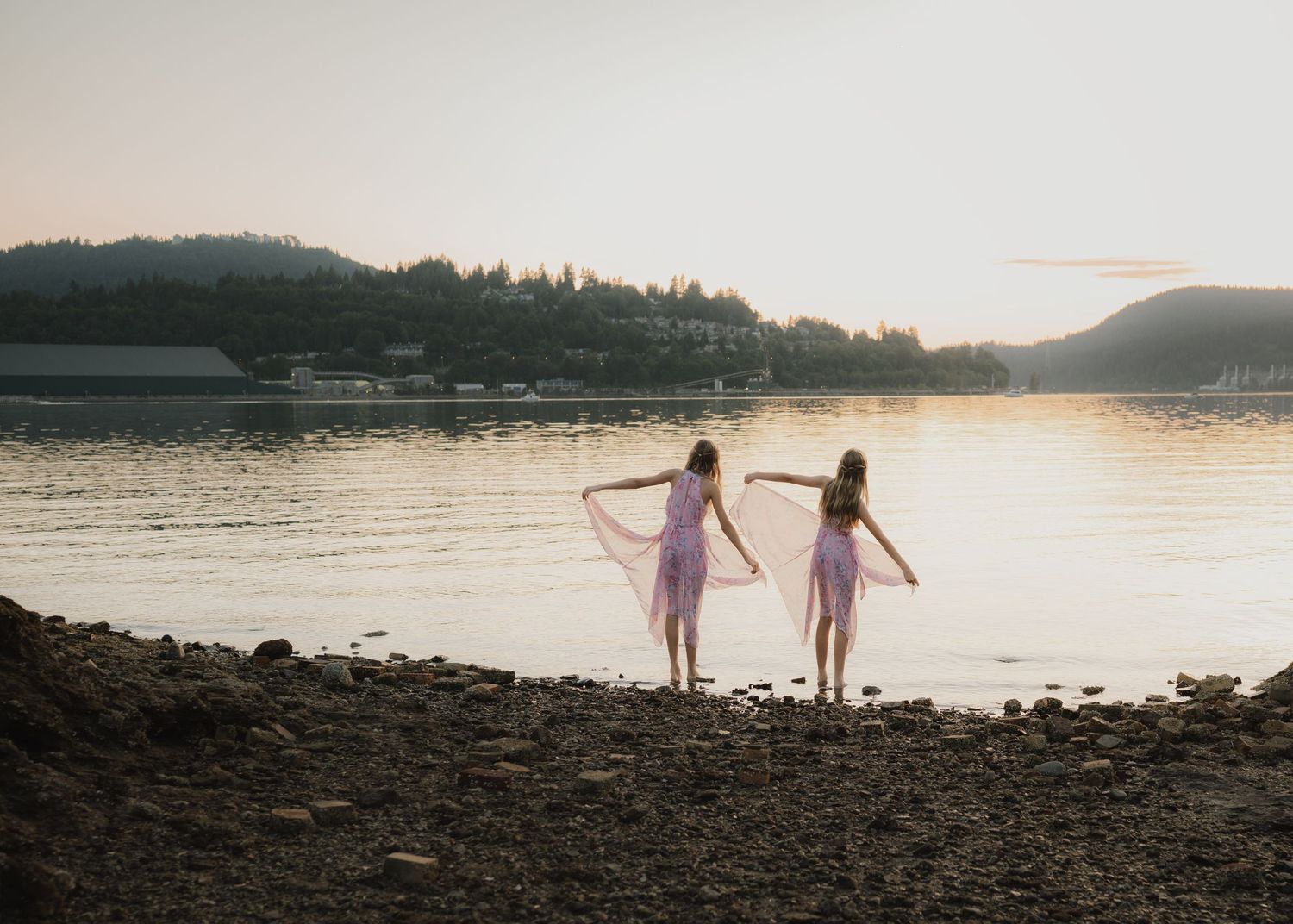 Two figures in pink dresses stand by the lakeside at sunset, silhouetted against the tranquil water and mountain landscape.