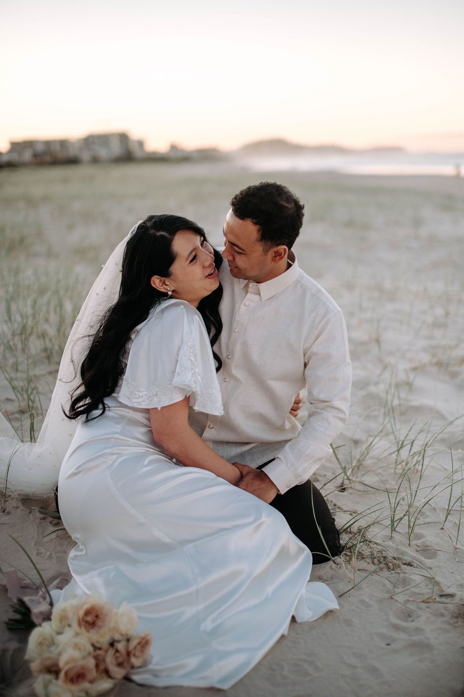 Romantic couple shares intimate moment on sandy beach at sunset with bouquet of roses nearby.