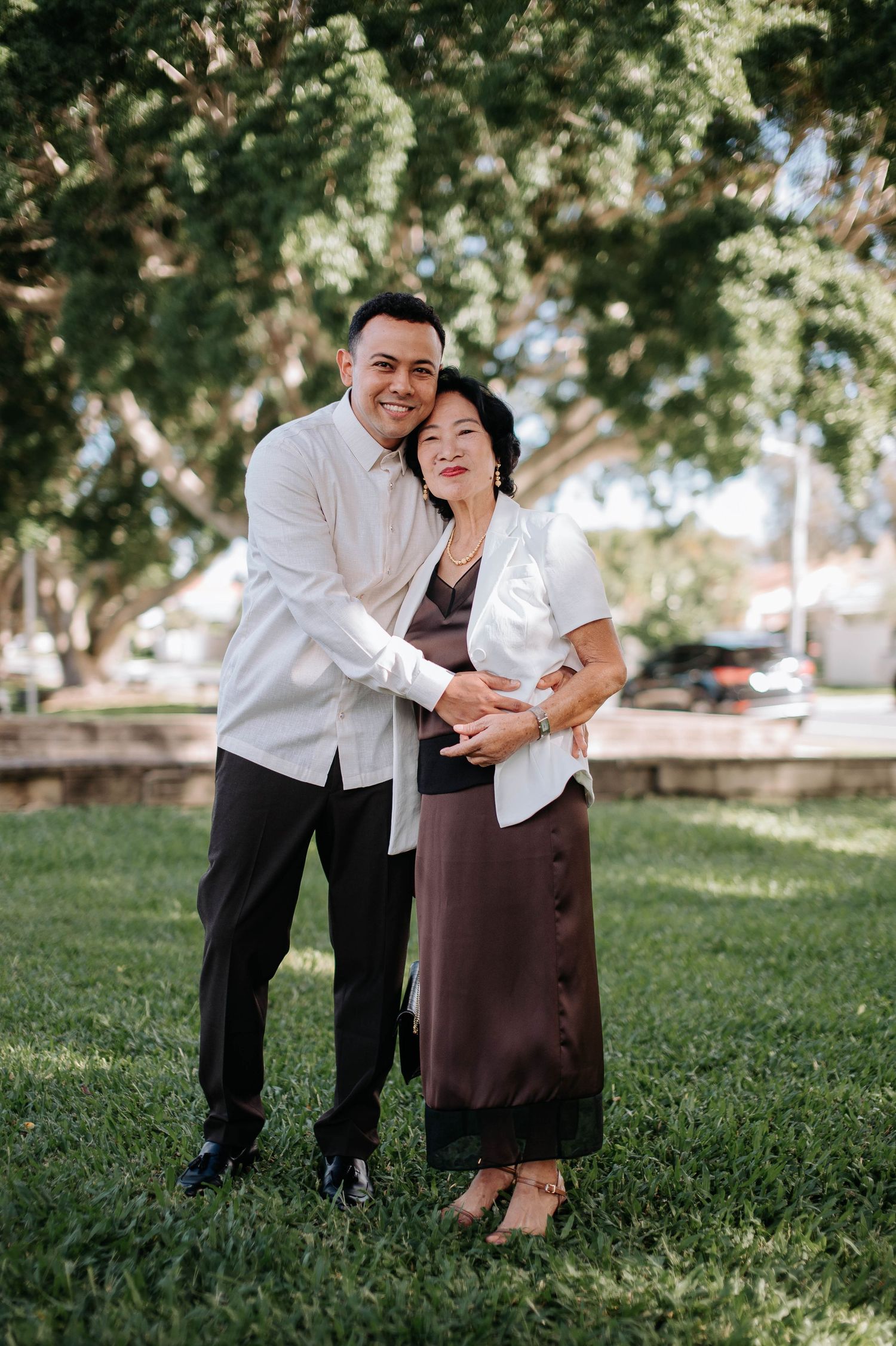 A couple embraces while standing in a park on a sunny day with trees in the background.