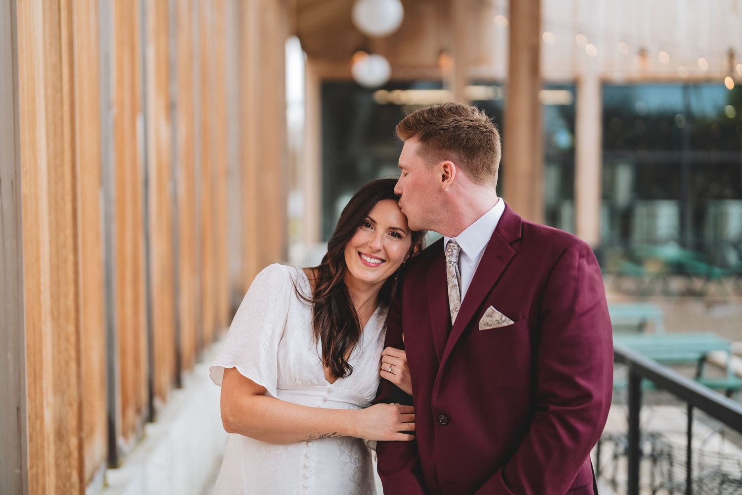 A couple shares a tender moment on their wedding day, with the groom in a burgundy suit and bride in white dress.