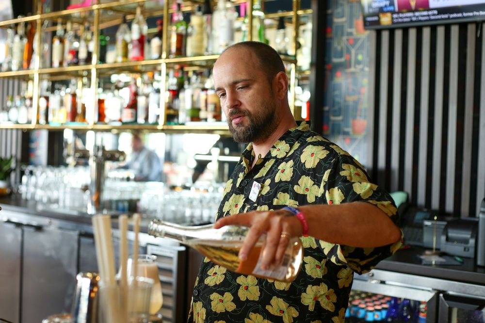 Bartender in a floral print shirt stands behind bar counter with shelves of liquor bottles in background.