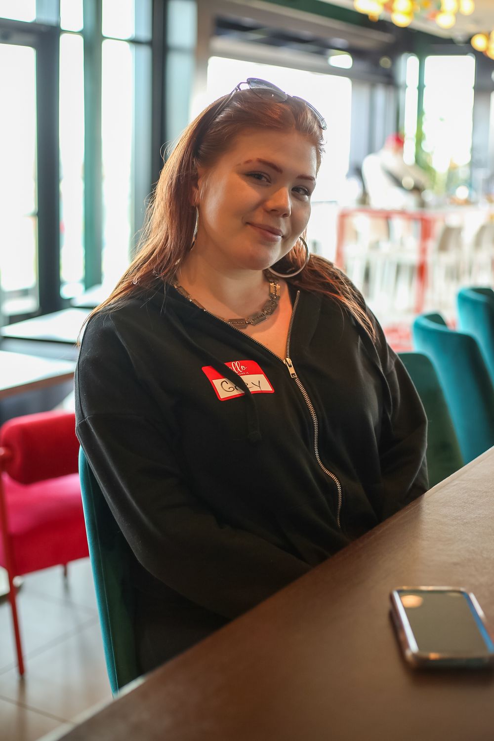 A person wearing a black zip-up hoodie with red patches sits at a table in a restaurant with turquoise chairs visible.