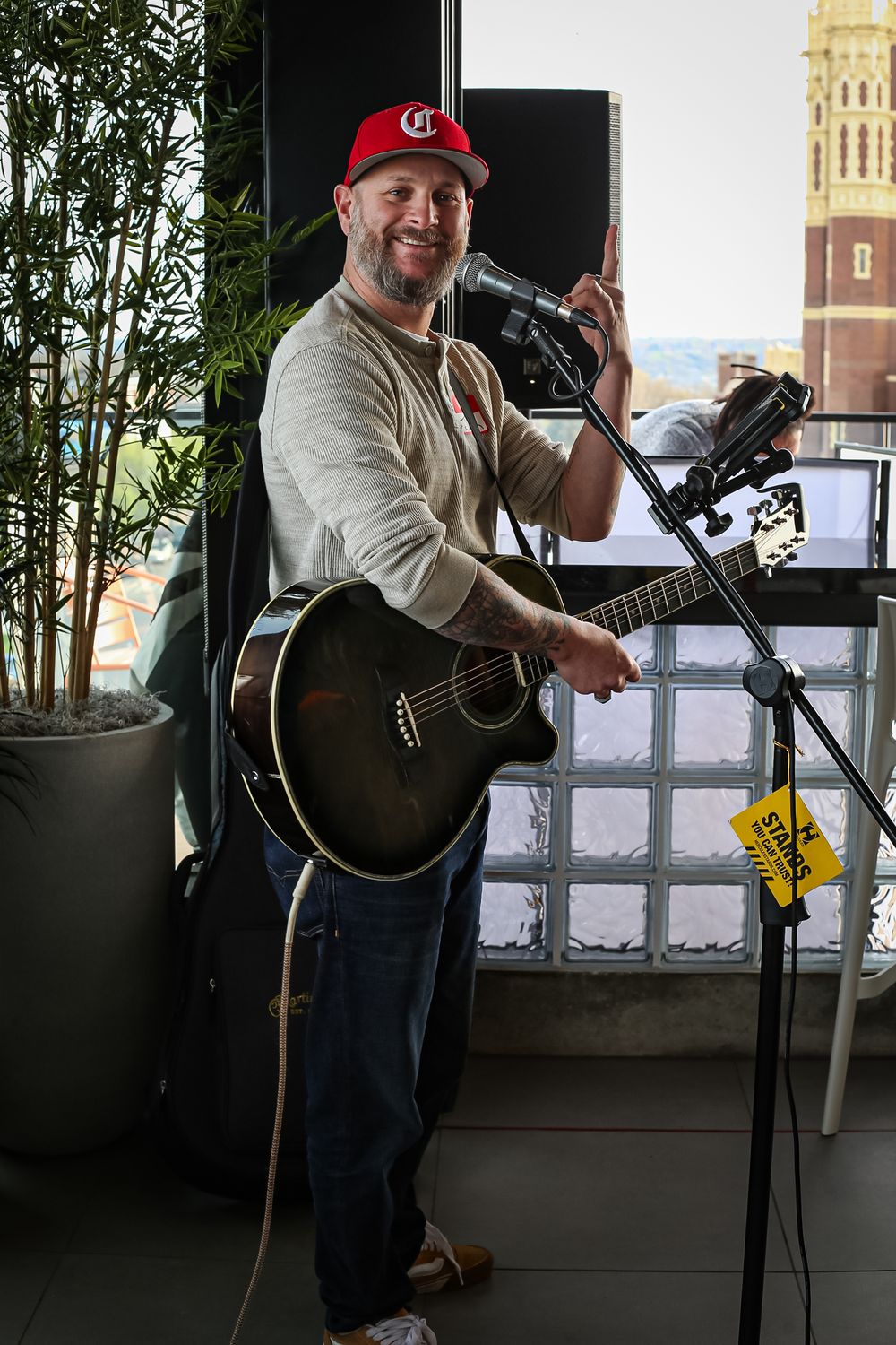 Musician performs with an acoustic guitar and microphone on a rooftop venue with city architecture visible in background.
