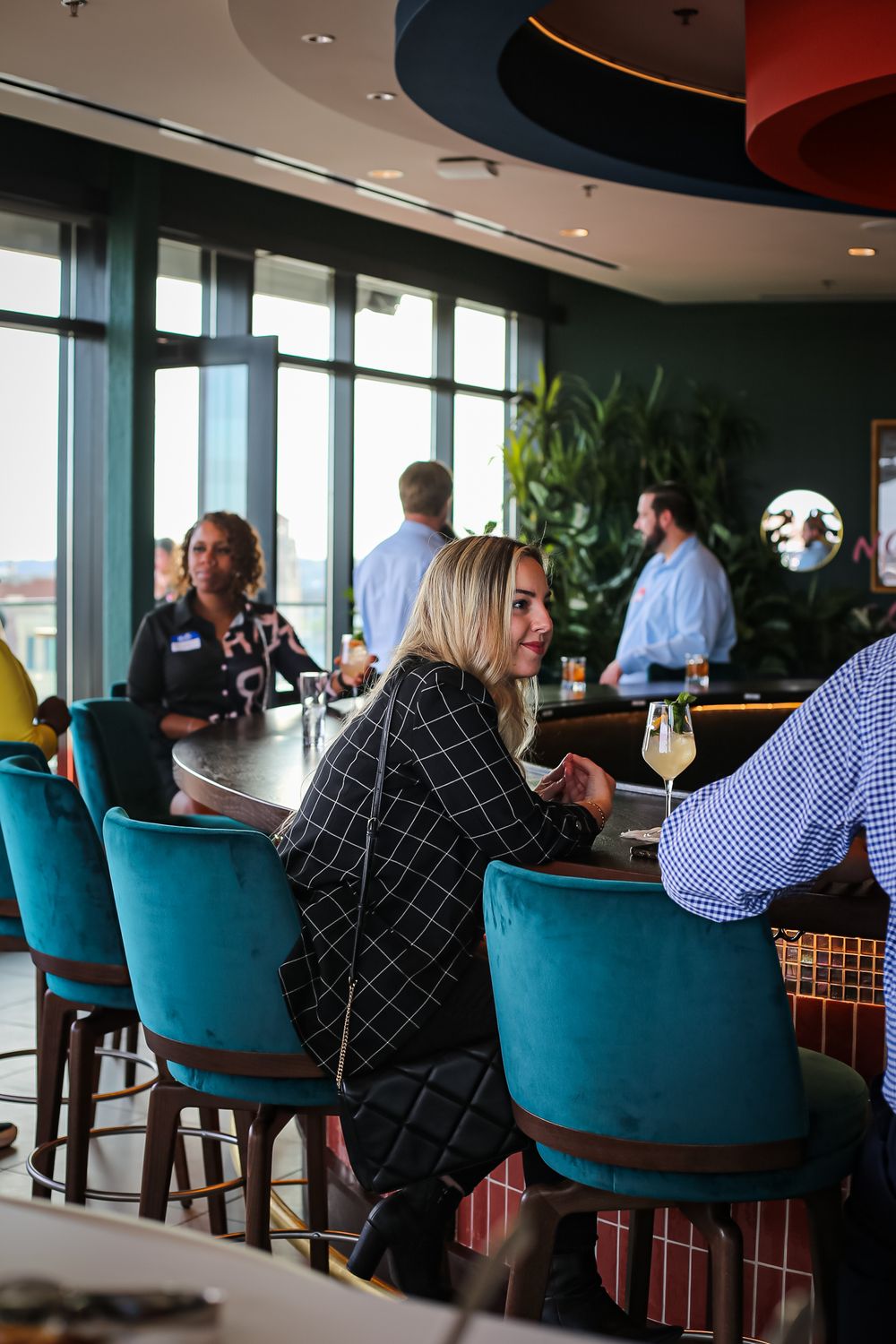 Restaurant patrons enjoy drinks at a modern bar with turquoise chairs and floor-to-ceiling windows.