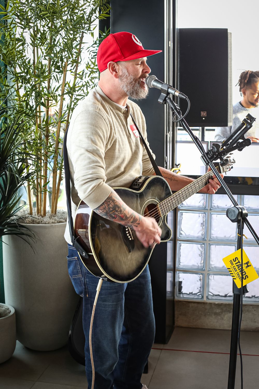 A performer in a red baseball cap plays acoustic guitar and sings into a microphone near potted bamboo plants.