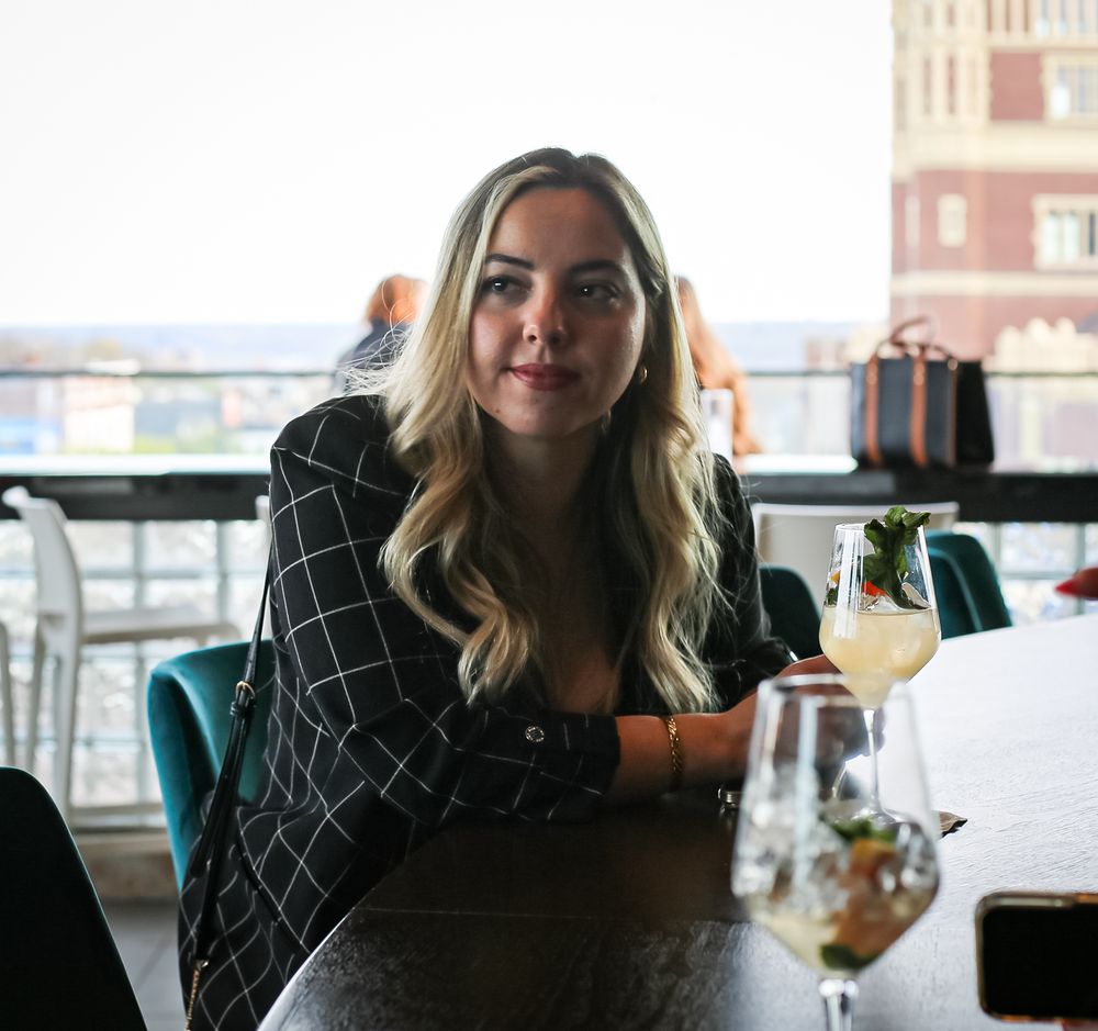 Woman in black patterned top sits at restaurant table with cocktail near window during evening dining service.